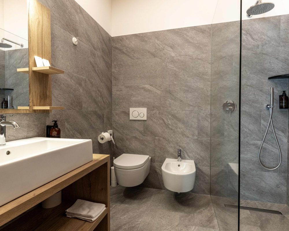 Interno del bagno moderno dello Chalet Alaska con pareti e pavimento in piastrelle grigie effetto pietra, box doccia in vetro, lavabo da appoggio su mobile in legno, WC e bidet sospesi.