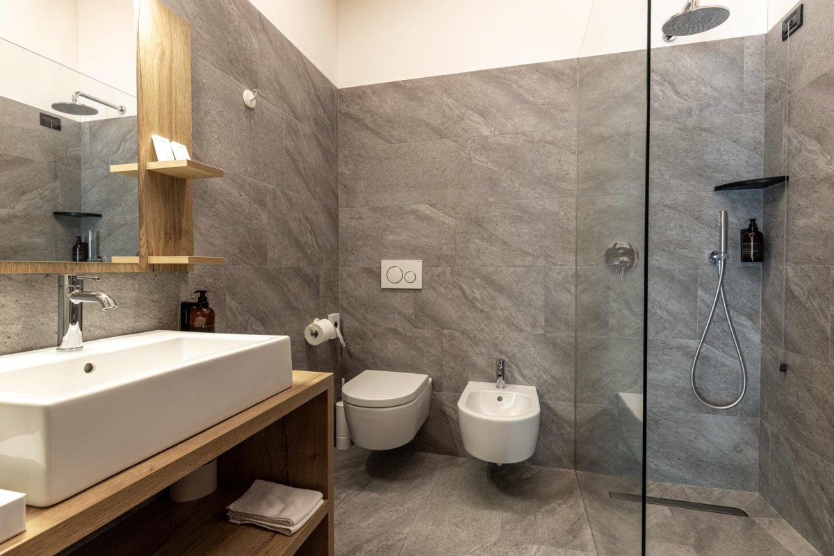 Interno del bagno moderno dello Chalet Alaska con pareti e pavimento in piastrelle grigie effetto pietra, box doccia in vetro, lavabo da appoggio su mobile in legno, WC e bidet sospesi.