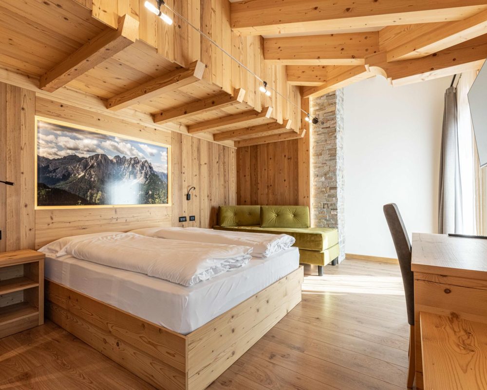 Moderna camera da letto matrimoniale in legno dello Chalet Alaska con testiera illuminata che incornicia un murale di montagne dolomitiche, scrivania e sedia.