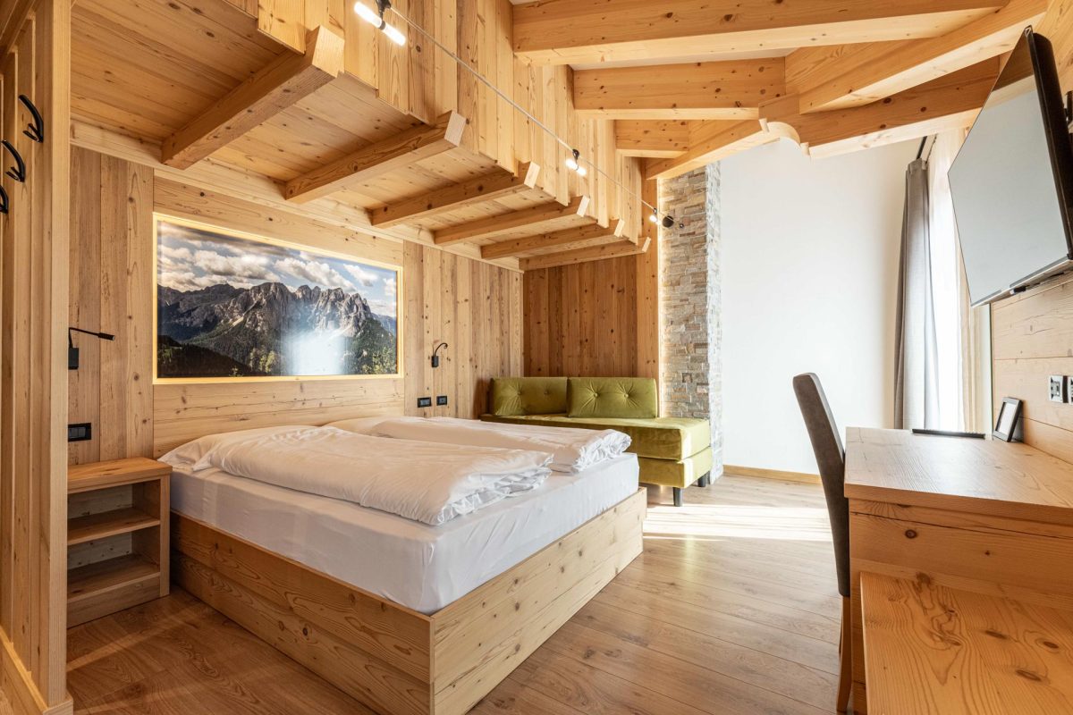 Moderna camera da letto matrimoniale in legno dello Chalet Alaska con testiera illuminata che incornicia un murale di montagne dolomitiche, scrivania e sedia.