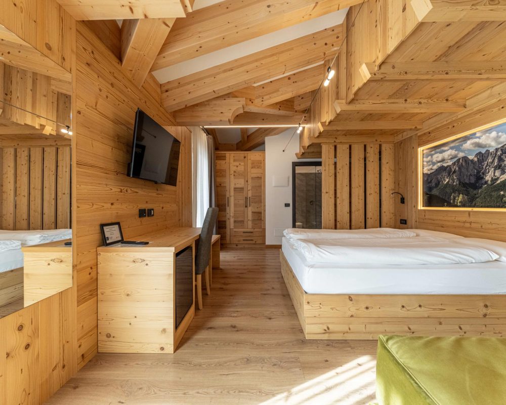 Ampia veduta della camera da letto dello Chalet Alaska, mostrando il murale retroilluminato sopra il letto matrimoniale, il divano letto verde oliva, la TV a parete e la scrivania con specchio.