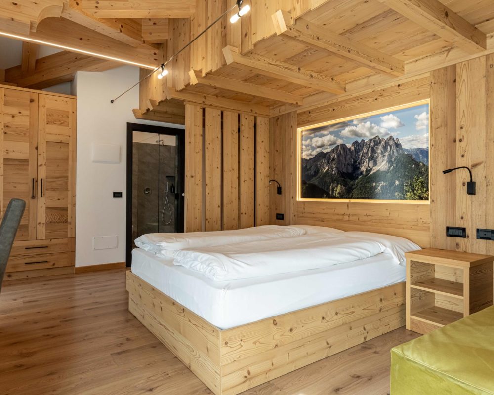 Veduta prospettica della camera dello Chalet Alaska, che si estende dalla scrivania al letto matrimoniale con murale, fino al bagno moderno con porta a vetro parzialmente visibile in fondo.