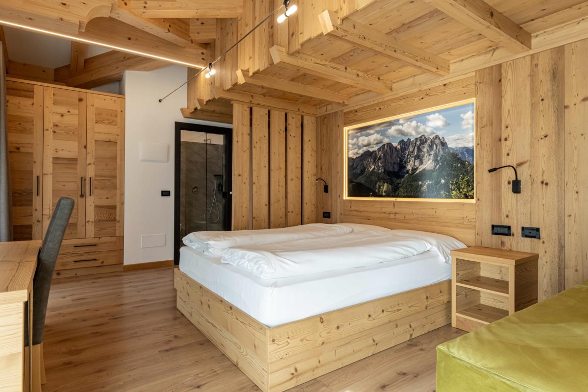 Veduta prospettica della camera dello Chalet Alaska, che si estende dalla scrivania al letto matrimoniale con murale, fino al bagno moderno con porta a vetro parzialmente visibile in fondo.