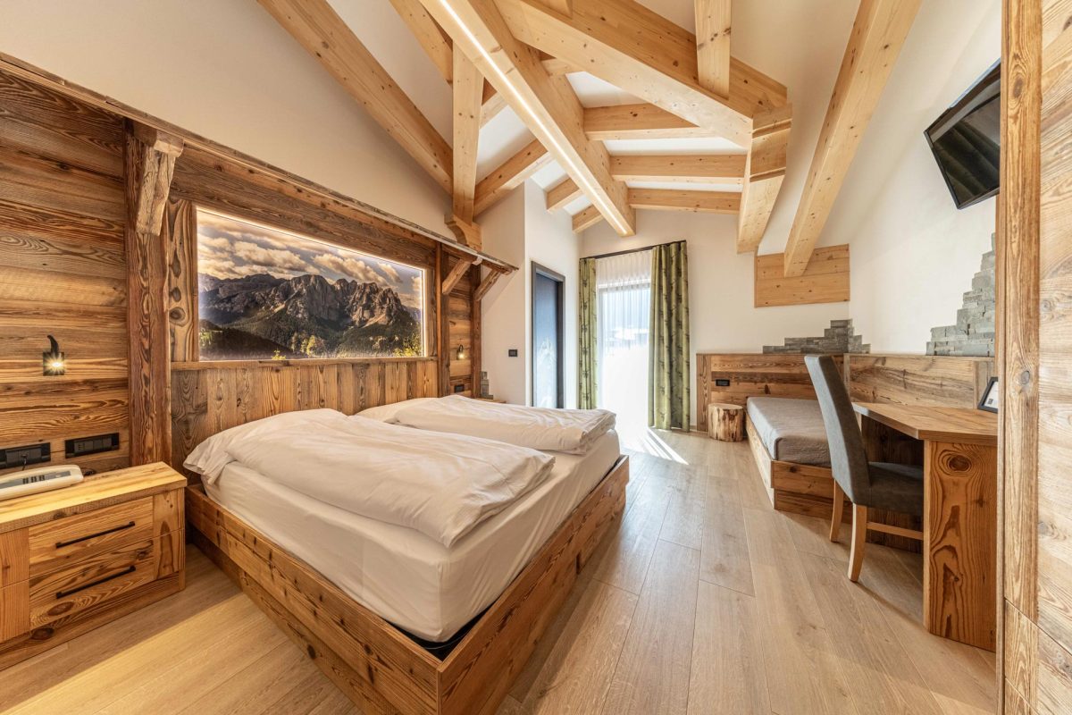 Camera da letto matrimoniale in legno in stile alpino moderno con travi a vista e grande quadro retroilluminato delle Dolomiti.